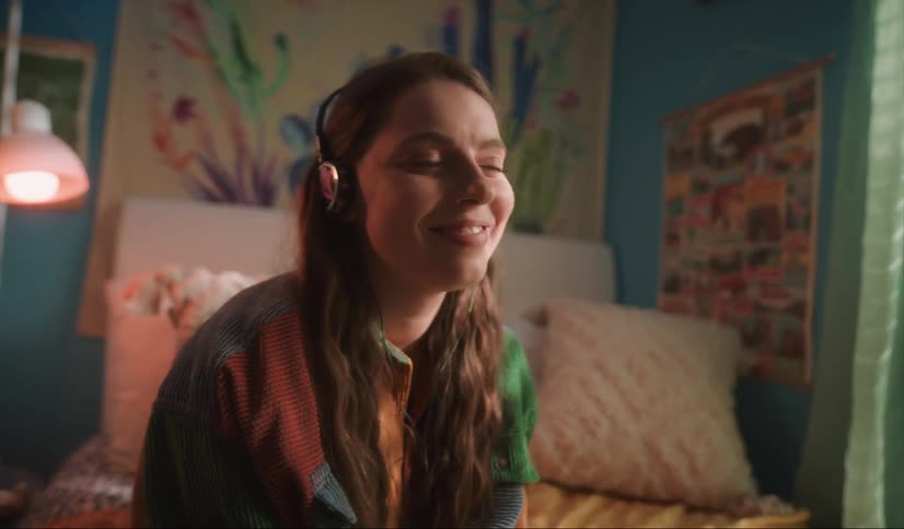 A teenage girl is sitting on her bed and smiling as she listens to her headphones