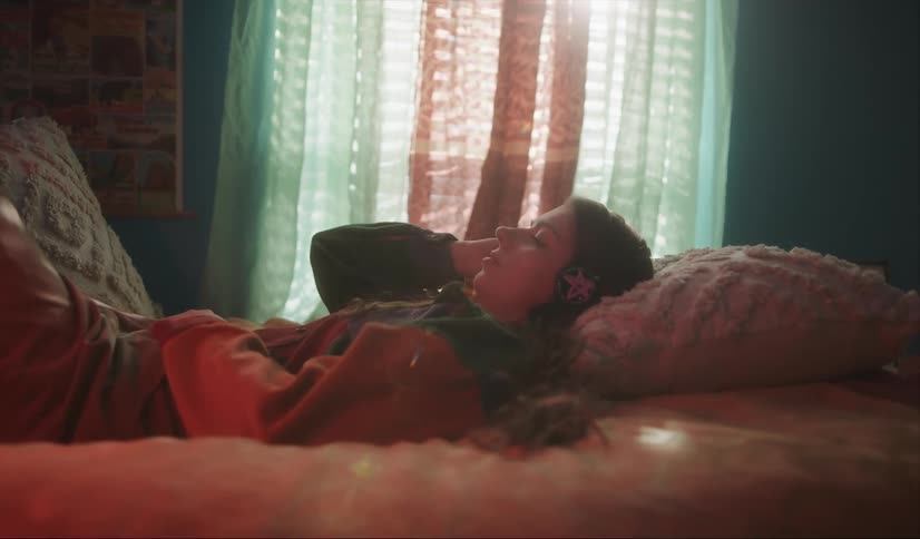 Girl with wavy hair colorful sweater and brown pants lies on bed listening to music with headphones in room