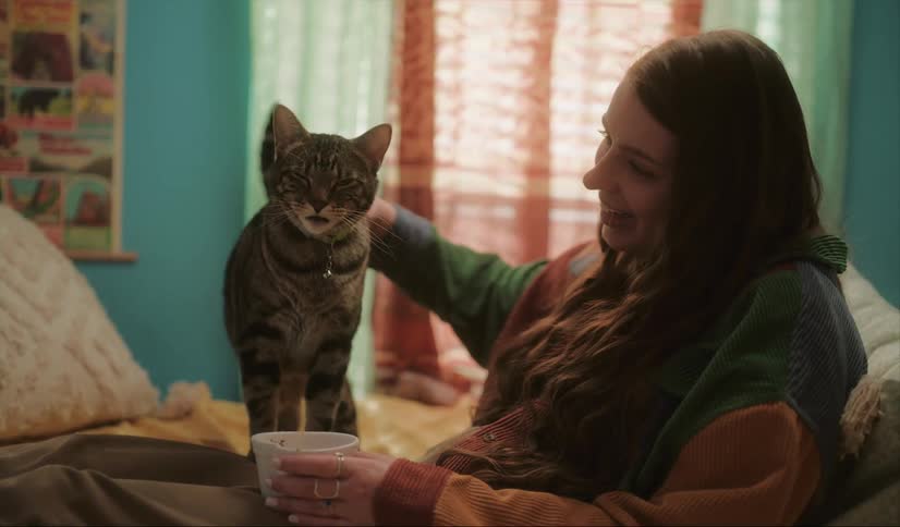 Woman with wavy hair and green sweater pets and feeds cat holding a bowl in room