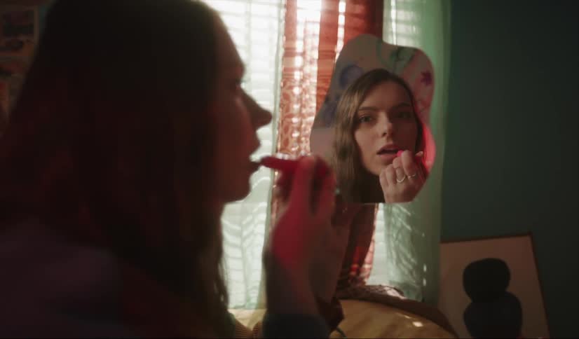 A teenage girl holds a mirror as she applies lipstick and then smiles
