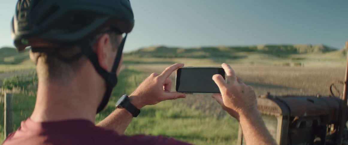 A man wearing a bicycle helmet stands outdoors and takes a photo of an old farm tractor