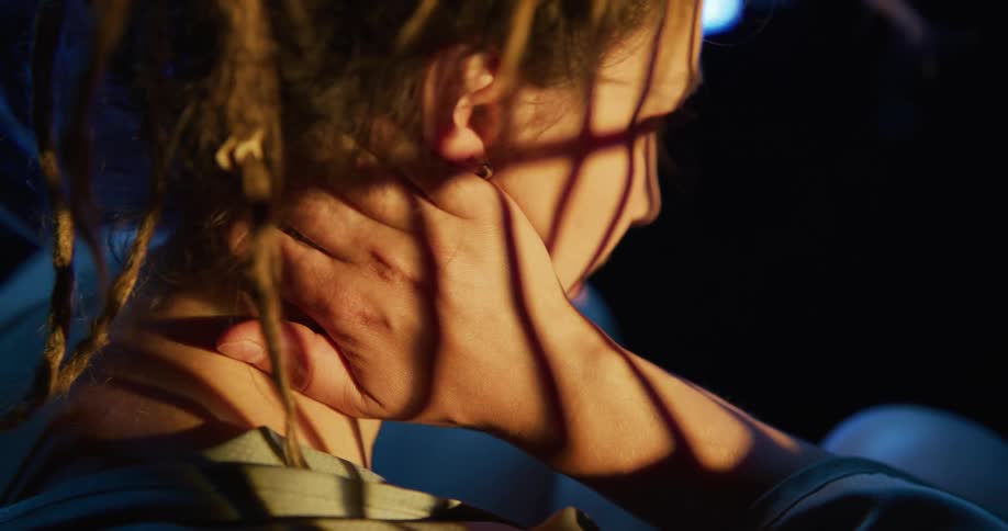 Young woman sitting inside a dimly lit room rubs her neck with her hand