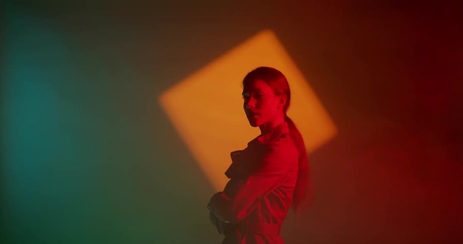Female model with ponytail and brown overcoat stands cross armed in spotlight