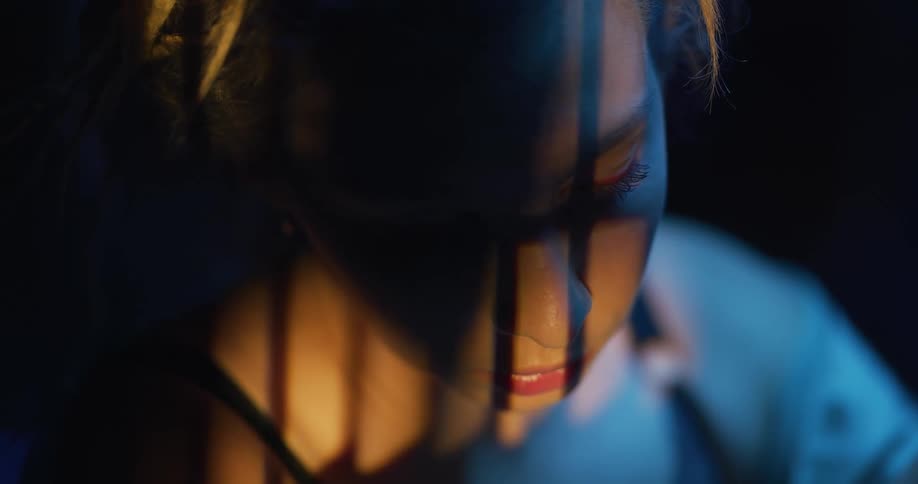 A woman sitting in the dark with light and shadows creating lines on her face rocks and sways
