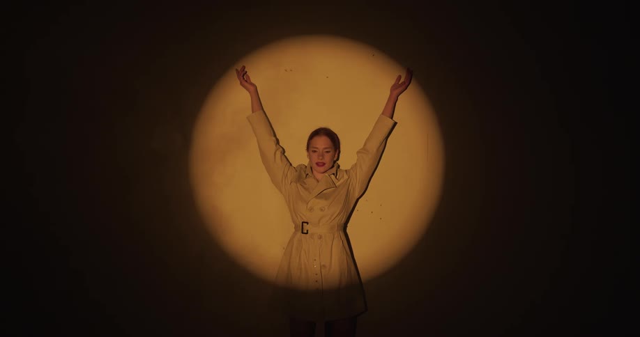 Female model with ponytail and brown coat stands in spotlight spreading and lifting arms up