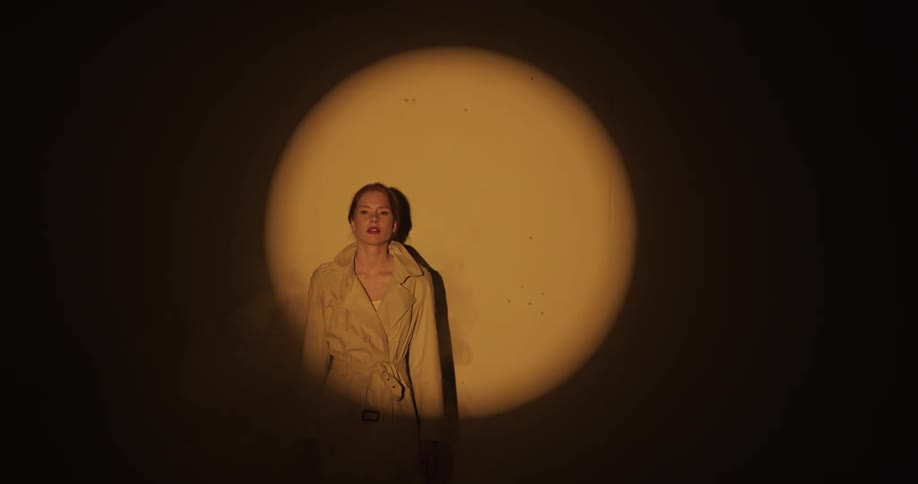 A woman is standing against a wall with a spotlight shining upon her as she poses for the camera