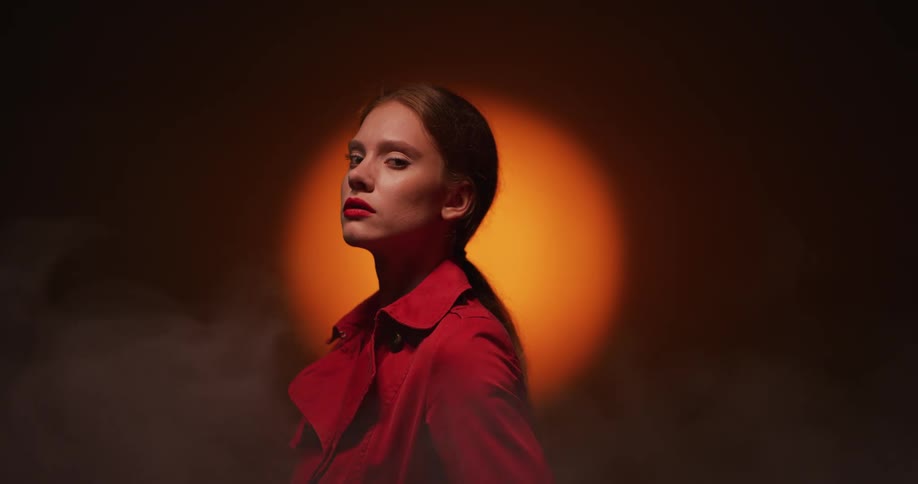 Female model with ponytail and brown overcoat stands in spotlight in room