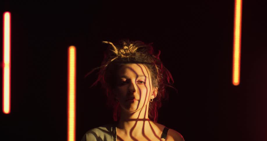 Young woman with a chaotic hairdo stands on a room surrounded by floating lights looking at the camera