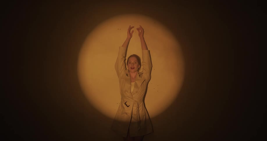 Jacket wearing woman illuminated by a spotlight raises her arms to clasp her hands together then place them on her head