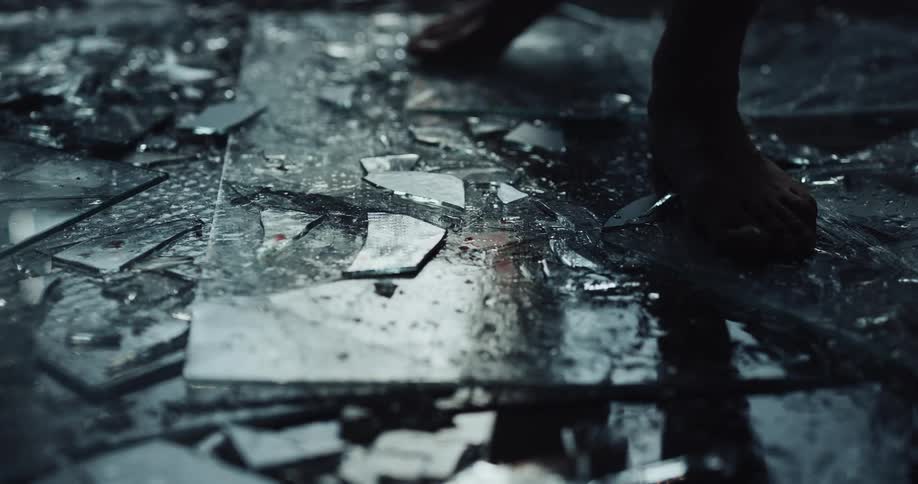 Barefoot person walks over wet broken pieces of glass on the floor of a dimly lit room