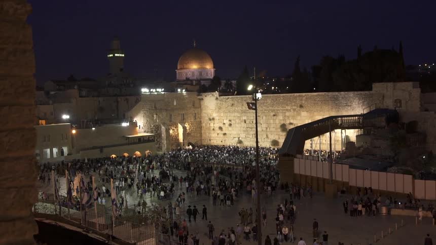 A crowd is standing before a stone wall in a square at night