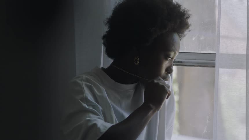 Woman with curly hair earrings and white shirt looks out the window and fiddles with necklace