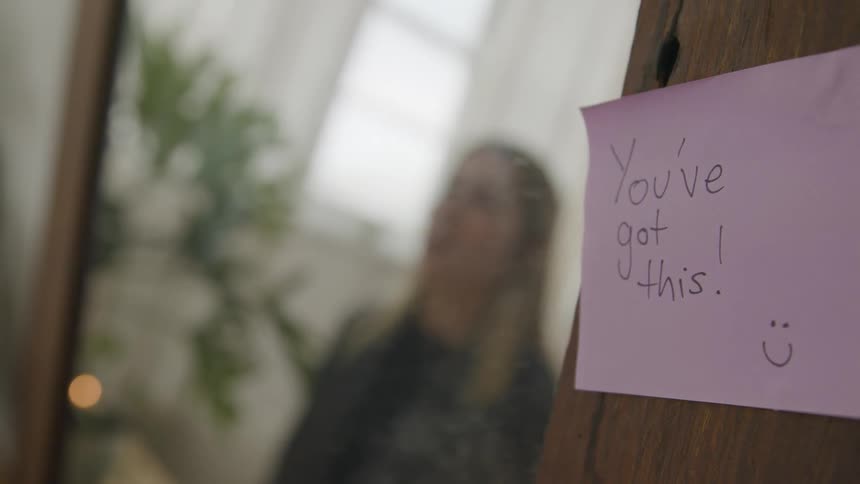 A woman looks at her reflection in a mirror that has a sticky note with encouraging words placed on it