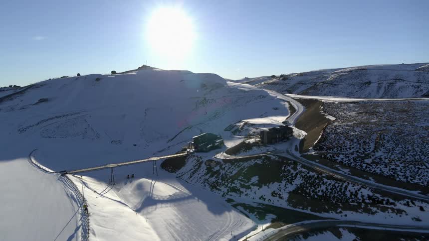 Aerial view of sun shining on snowy mountain with roads leading to building next to bridge over frozen river