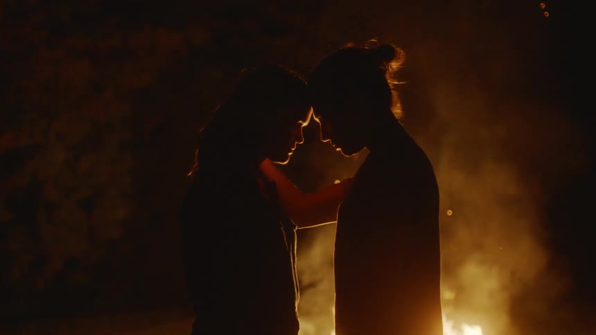 A man and woman stand close together in the darkness in front of a bonfire