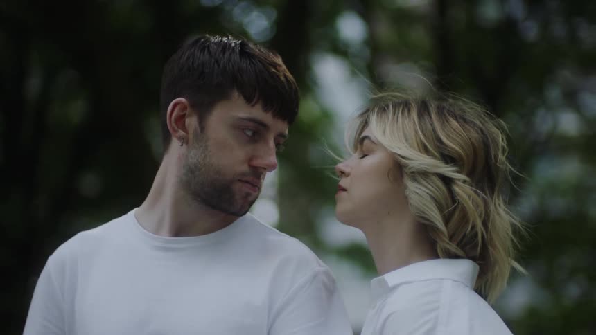 Woman with fair hair and white coat cuddles man with beard earrings and white t shirt in the forest