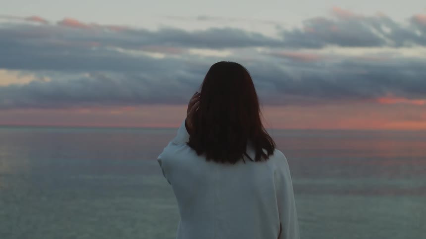 Young woman wearing a white sweater stands in front of the ocean at sunset
