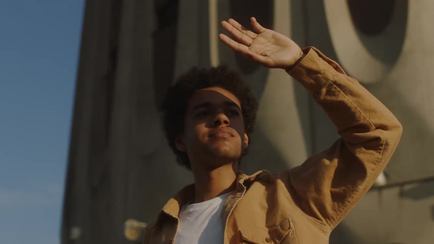 Young black man standing outdoors at a sunny day looking ahead and covering his face against the sunlight with his hand