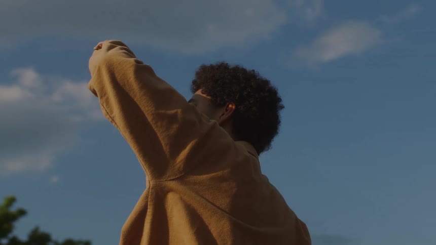 Young black man standing outdoors looking ahead with his arm covering his face against the sunlight