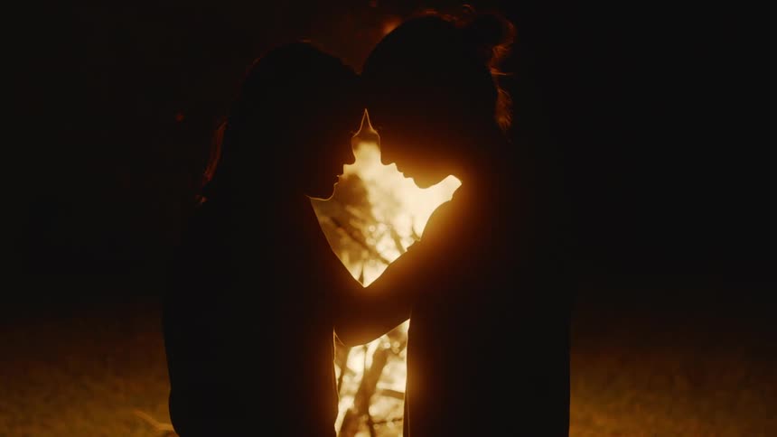 Silhouettes ofa young couple standing in front of a camp fire at night touching heads and looking at each other romantically