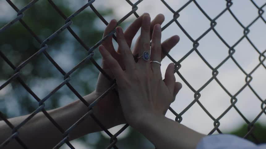 Two people clasp hands from either side of a chain link fence
