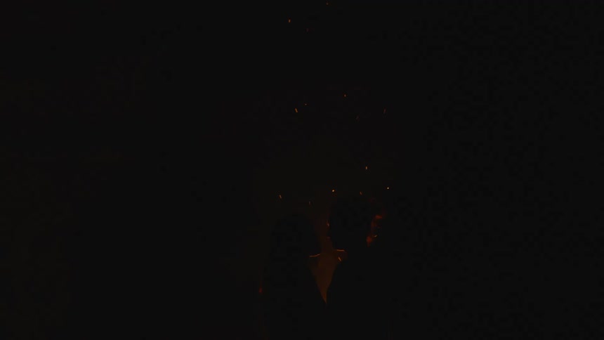 A couple are outside kissing near a bonfire as sparks rise in the darkness
