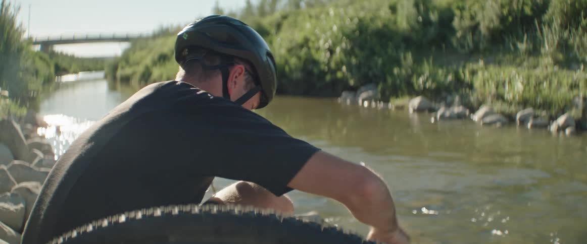 A man wearing a helmet sits with his bicycle next to a river