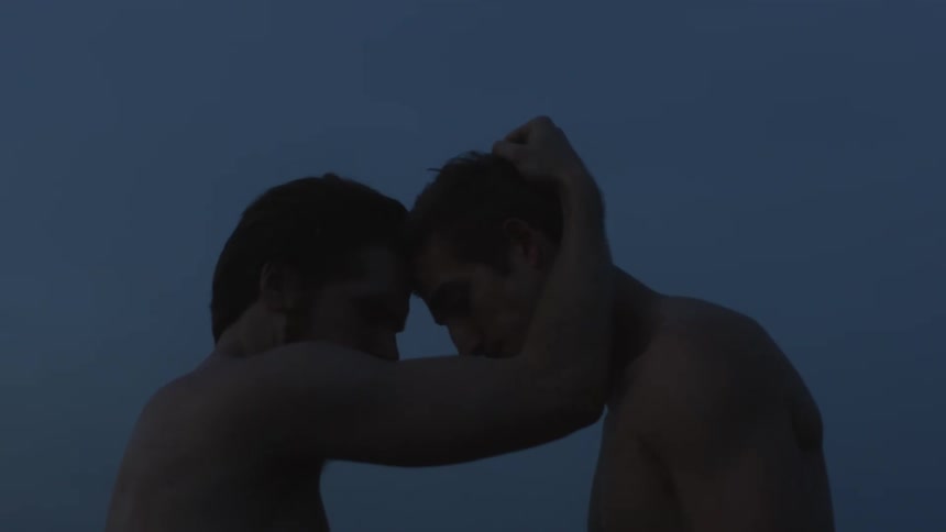 One man grasps the hair of another man and brings their foreheads together while they stand outdoors