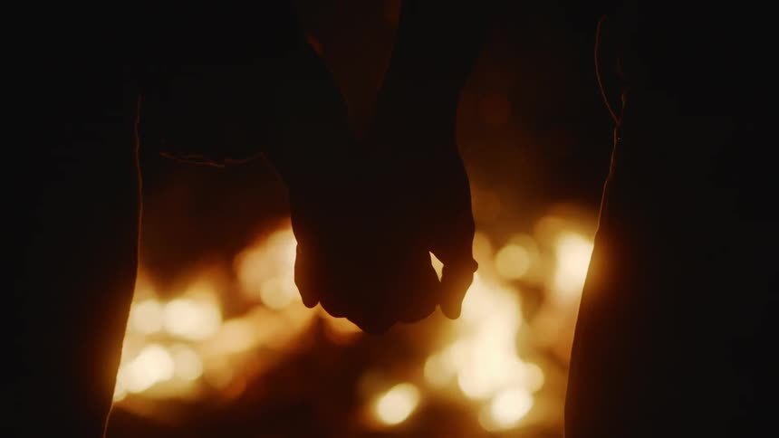 Silhouette of a couple holding hands in front of a large bonfire at night