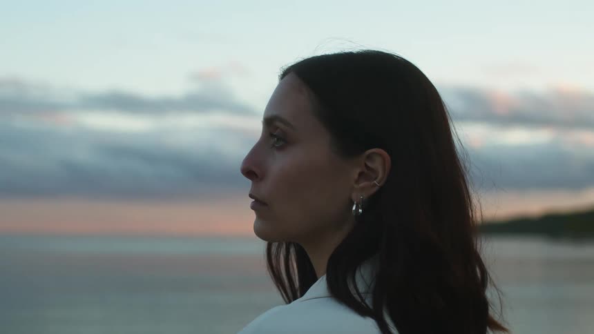 Woman with wavy hair earrings and white jacket looks at sea on the beach at sunset