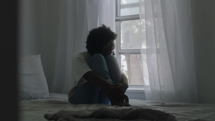 Thoughtful and sad black woman sitting on bed next to the window looking outside