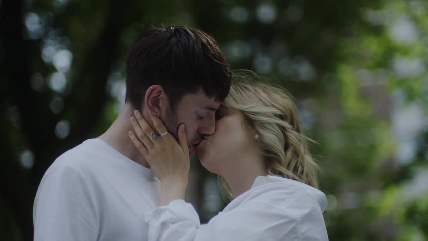 Man with beard and white shirt makes out with blonde woman wearing white coat and rings in the forest