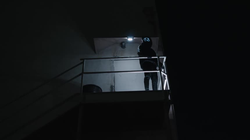 Person wearing a digital mask stands at top of staircase illuminated by electric light and surrounded by darkness