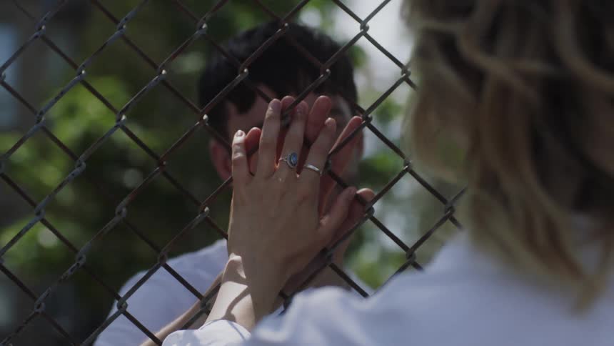 Young couple look at each other and touch hands on opposite sides of a chain link fence