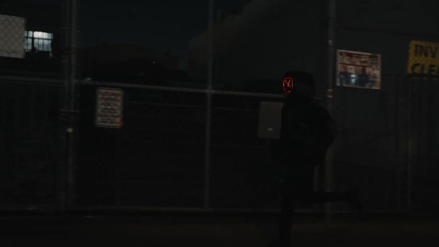 A person with a glowing mask on runs past stores and parked vehicles at night