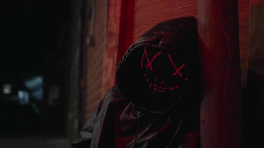 Image on mask that hooded person is wearing lights up as the person leans against building at night
