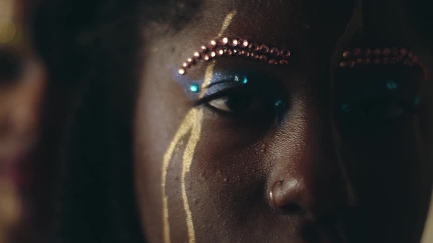 A woman is wearing colorful beaded eye makeup and there is another woman standing behind her