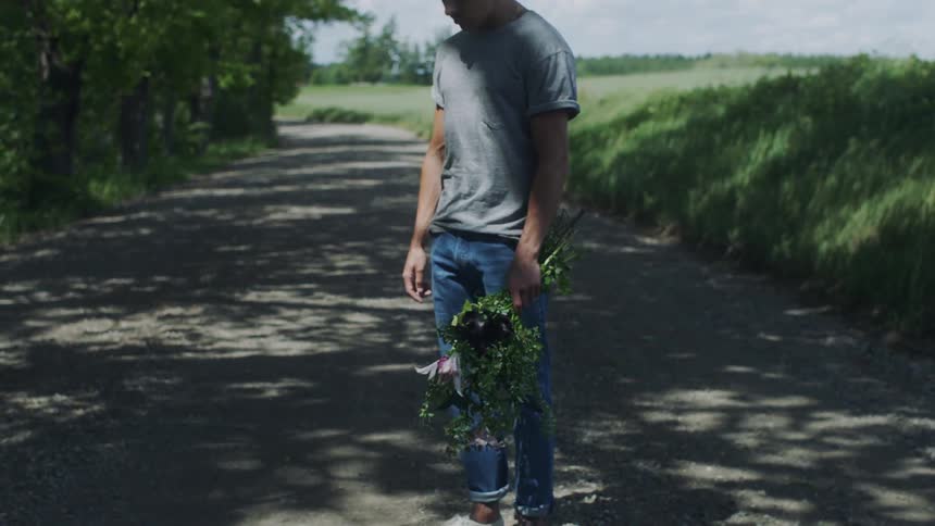 Disappointed man with bun gray shirt jeans and white sneakers holds a bunch of flowers standing in the middle of country road in the countryside