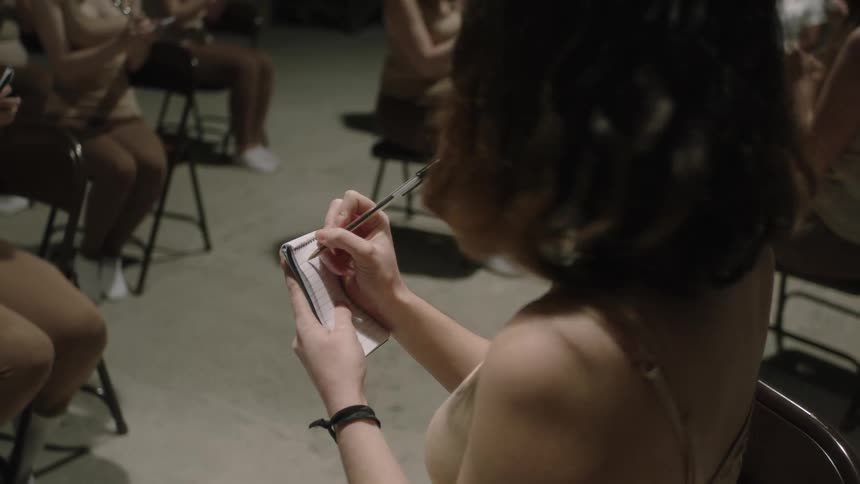 A woman with dark hair is standing and writing on a notepad in her hand as other women are sitting in chairs