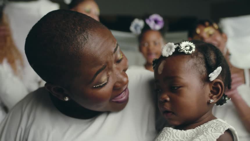 A black woman wearing white clothes puts a mark on a toddler forehead using her thumb in front of a crowd