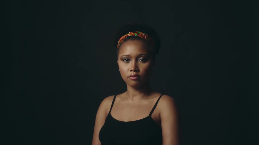 Young black woman sitting on a room in front of a black background turns around to look at the camera
