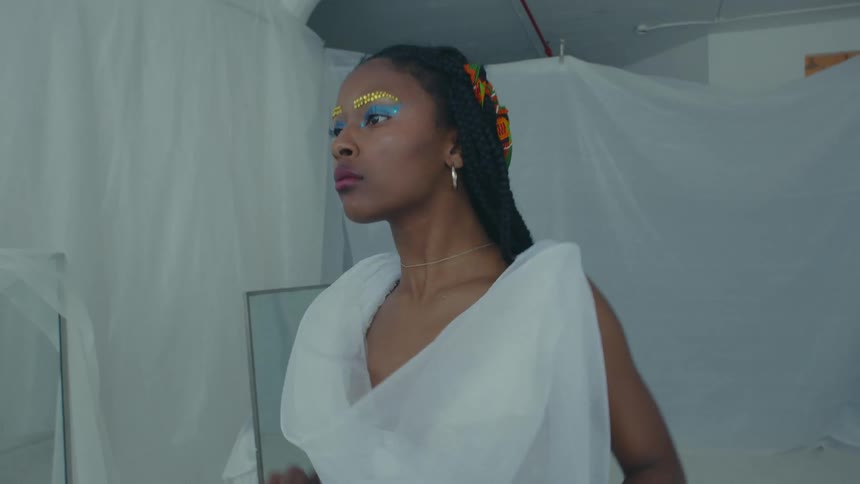 A woman wearing white standing in a plastic sheeted room with mirrors begins to dance