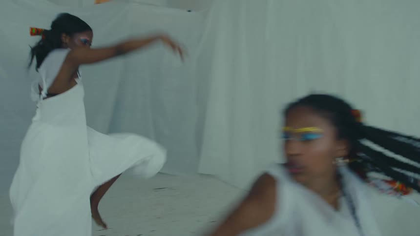 Three women wearing white dresses dance and perform in a room covered in white fabric