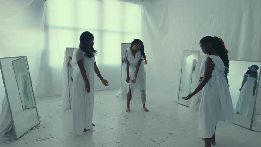 Three women dressed in white are dancing near mirrors in a room covered with white sheets