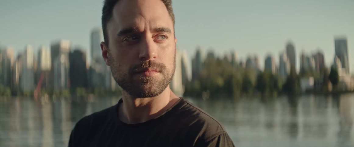 Man with black t shirt and beard looks over his shoulder next to sea with city skyline in the background