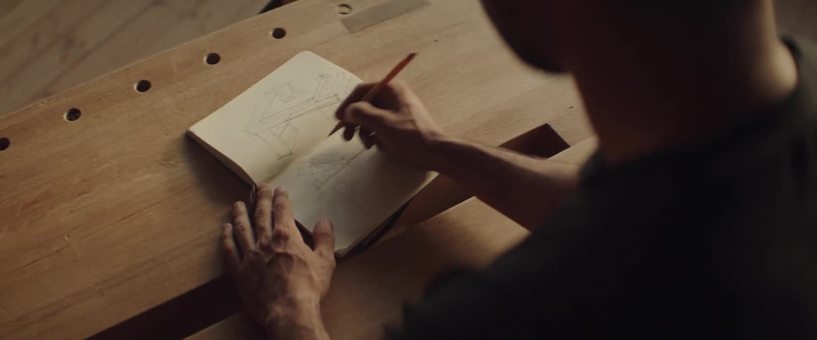 Man sketching in a notebook with a pencil on a wooden bench