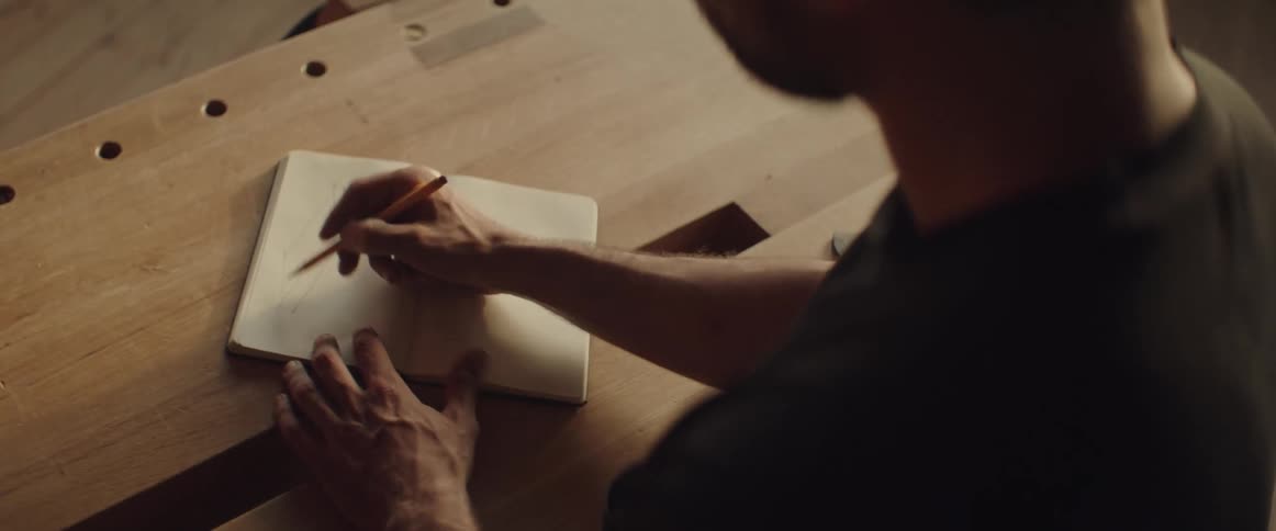 Man standing in front of a wood table drawing on his notebook with a pencil