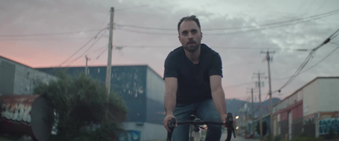Man with beard black t shirt and jeans rides bicycle on road in the neighborhood at sunset