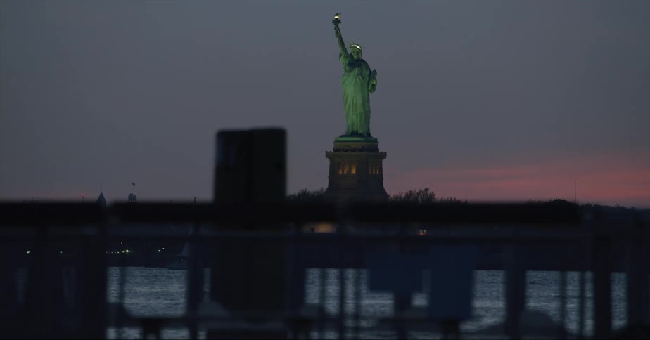 A person is walking near the water as the statue of liberty stands tall overlooking new york harbor