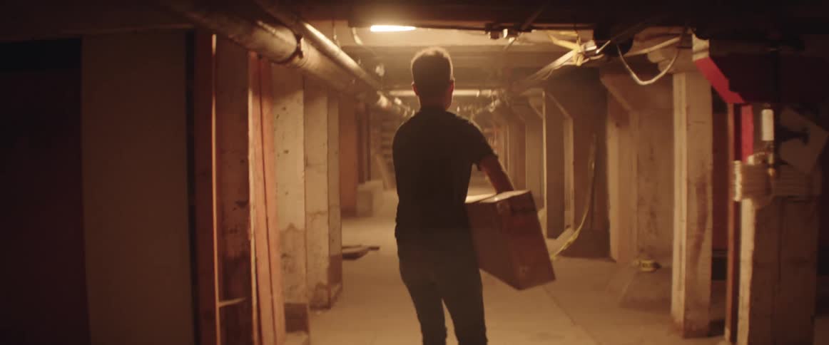 A man carried a heavy piece of lumber through a low ceilinged basement corridor